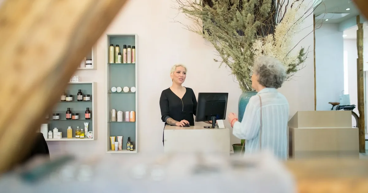 Eine Kundin spricht am Empfangstresen eines hellen Friseursalons mit einer Friseurin, die am Bildschirm den Termin aufruft.