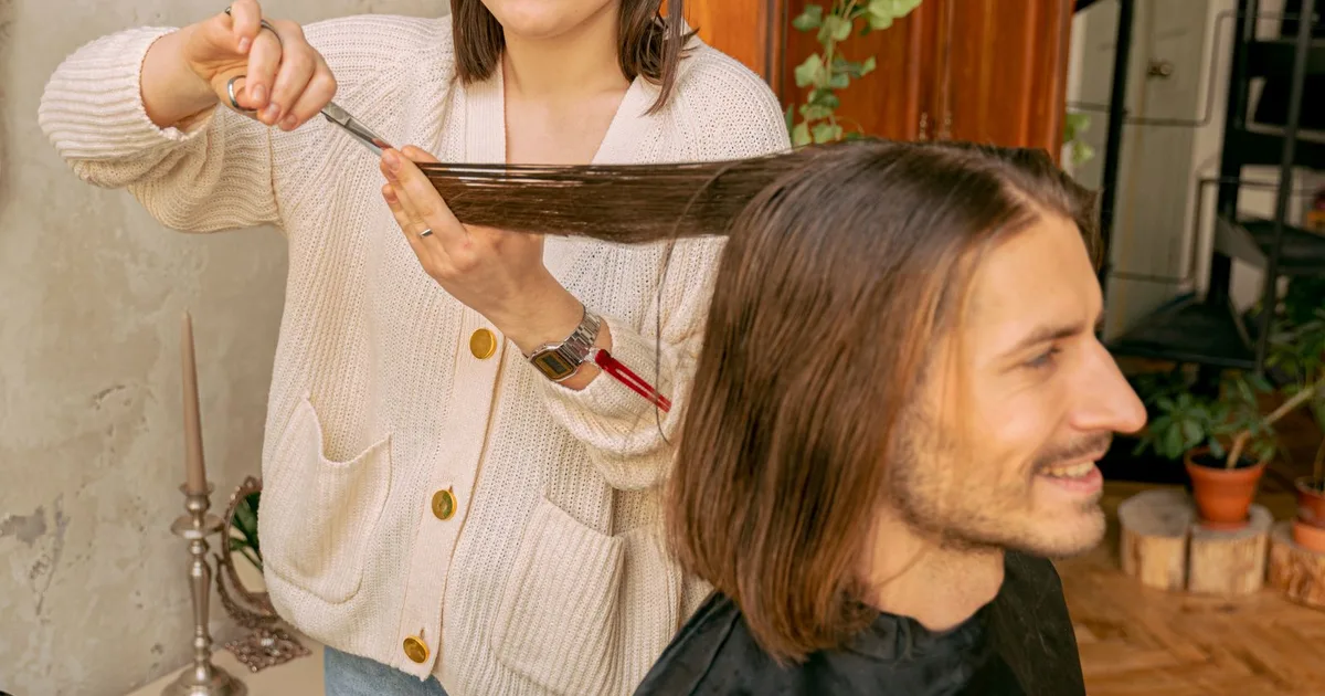 Eine Friseurin schneidet das lange braune Haar eines Mannes in einem hellen Raum mit Pflanzen und warmen Holzmöbeln.